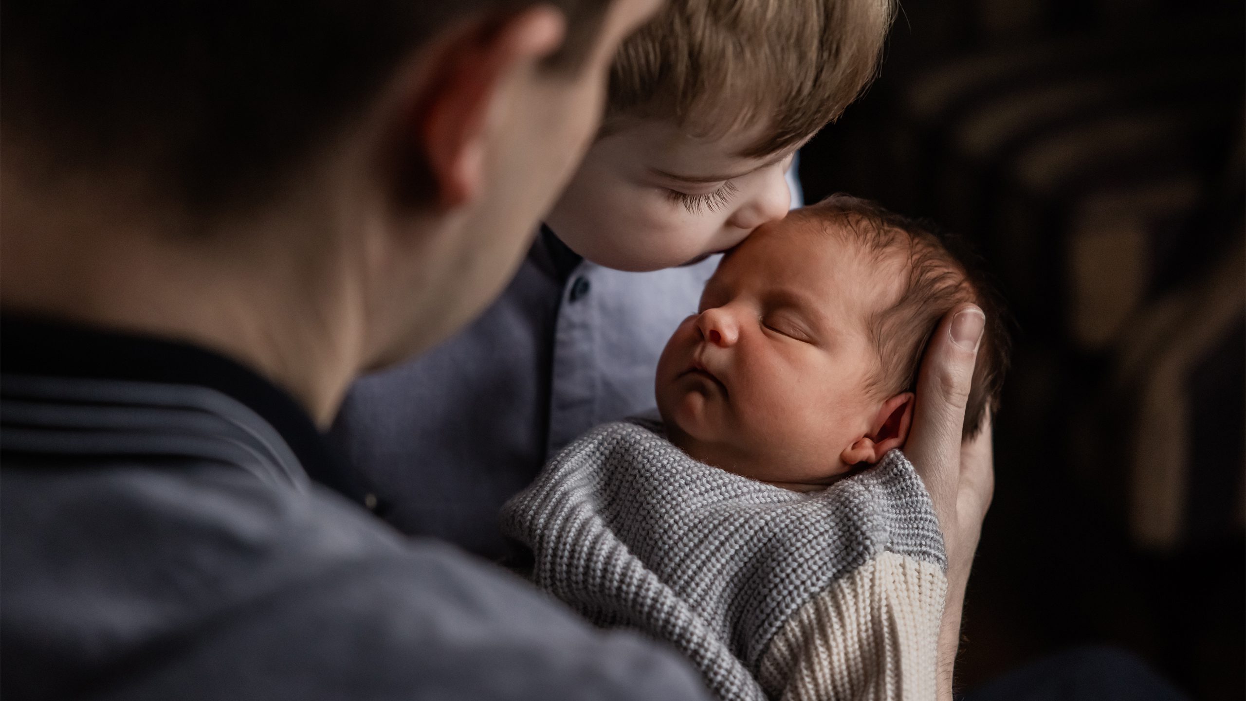 View over dad's shoulder of a boy kissing his newborn sister's forehead.