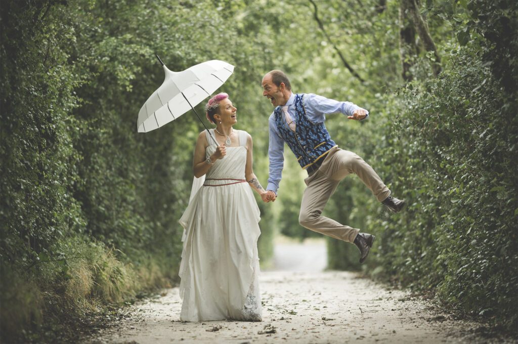 milca-and-wedding-photography-angela-cunning Wedding couple on a light gravel path surrounded by trees. The bride has pink hair and is holding an umbrella, the groom is up in the air, clicking his heels together.